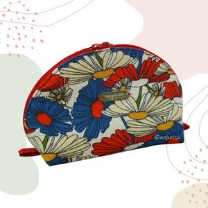 Könnte beinhalten: Eine halbmondförmige Kosmetiktasche mit rotem Reißverschluss und Anhänger. Die Tasche hat ein Blumenmuster aus weißen, roten und blauen Gänseblümchen auf cremefarbenem Hintergrund. Der Markenname "anjurisa" ist auf einem kleinen goldenen Etikett zu sehen.