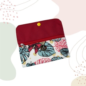 May include: A small, open wallet with a red flap and a zipper closure. The wallet features a tropical leaf pattern in blue, pink, and white. The interior is red, and the wallet is secured with a yellow snap.