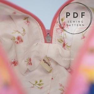 May include: A fabric pouch interior featuring a white lining with a pink floral pattern, a pink zipper closure, and a small beige fabric tag labeled anjurisa. The image includes a circular overlay with the text PDF SEWING PATTERN.