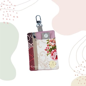 Peut inclure: Un porte-monnaie rectangulaire en tissu avec un motif floral sur fond blanc, une section latérale à imprimé géométrique rouge et marron foncé, une fermeture éclair beige, un panneau supérieur rose avec bouton-pression et un mousqueton en métal argenté.