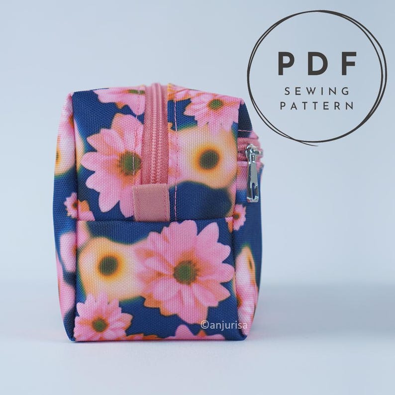 May include: A rectangular zippered pouch featuring a dark blue background with a repeating pattern of pink daisy-like flowers with yellow centers. The pouch has a pink zipper closure and a matching pink fabric pull tab. Text reads PDF SEWING PATTERN.