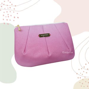 Può includere: Una pochette rosa glitterata con chiusura a zip, dotata di un cursore in metallo dorato e di una piccola targhetta rettangolare dorata centrata sul davanti con il testo anjurisa inciso in corsivo. La pochette presenta un design plissettato.