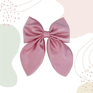 Pode incluir: Um laço de cabelo na cor rosa seco, feito de um tecido liso e acetinado. O laço apresenta duas grandes alças arredondadas no topo e duas longas pontas afuniladas que descem. O centro é preso com um nó do mesmo tecido.