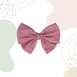 Puede incluir: Un lazo para el pelo de tela rosa empolvado con un nudo central y dos bucles anchos y simétricos plegados a cada lado. La tela tiene un aspecto mate y texturizado. El texto anjurisa está impreso en letras blancas pequeñas a la derecha.