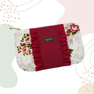 Op de afbeelding: Een rechthoekige clutch met bloemenprint, rode ruches en een gouden rits. De clutch heeft een crèmekleurige achtergrond met rode en roze rozen. Een gouden label met de tekst "anjurisa" is gecentreerd op het rode paneel.