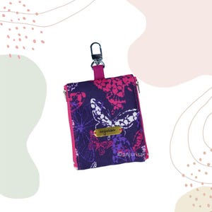May include: A small, zippered wallet with a butterfly pattern in shades of purple and pink. The wallet has a metal clasp and a small tag that reads "anjurisa". The zipper and strap are a bright pink color.