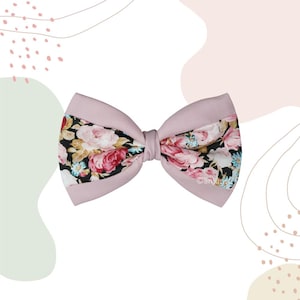 May include: A bow accessory featuring a solid light pink fabric border and a central panel with a black background printed with pink roses, white flowers, and small blue accents. The bow is tied in the centre with a matching light pink fabric knot.