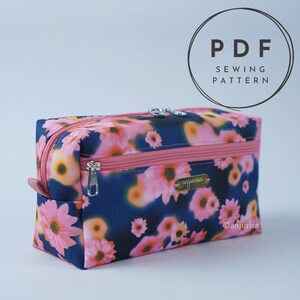 May include: A rectangular zippered pouch featuring a dark blue background with a pink and orange floral pattern. The pouch has a pink zipper, a front zippered pocket, and a small gold-toned metal label with the text anjurisa attached.