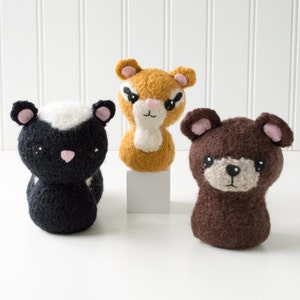 May include: Three crocheted stuffed animals: a black and white skunk, a brown bear, and a tan and white squirrel. The animals are all sitting on a white surface.