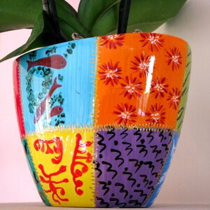 May include: A colourful ceramic planter with a patchwork design. The planter is decorated with flowers, fish, and abstract patterns in shades of blue, orange, yellow, green, and purple.