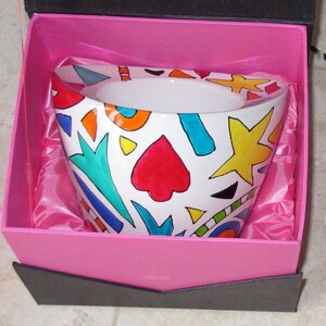 Può includere: Una ciotola in ceramica bianca con un colorato design astratto di cuori rossi, stelle gialle e forme blu, arancioni e verdi. La ciotola è in una confezione regalo rosa con fodera in raso rosa.