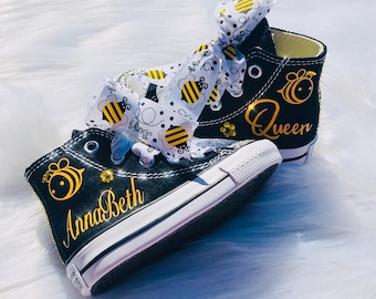 May include: Black high-top canvas shoes with a white ribbon bow and bee design. The shoes are personalized with the name "AnnaBeth" and the word "Queen".