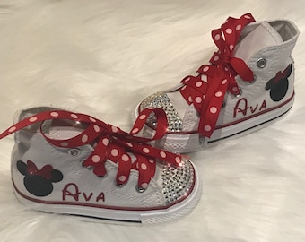 May include: White canvas high-top sneakers with red and white polka dot ribbon laces. The shoes have a black Minnie Mouse silhouette with a red bow on the side and the name "Ava" written in red. The shoes are decorated with rhinestones.