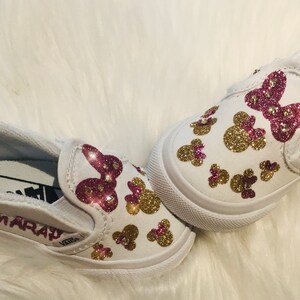 May include: A pair of white slip-on shoes decorated with pink and gold glitter. The shoes have a pink glitter bow and gold glitter Mickey Mouse heads. The left shoe has the name "Amaray" written on the side.