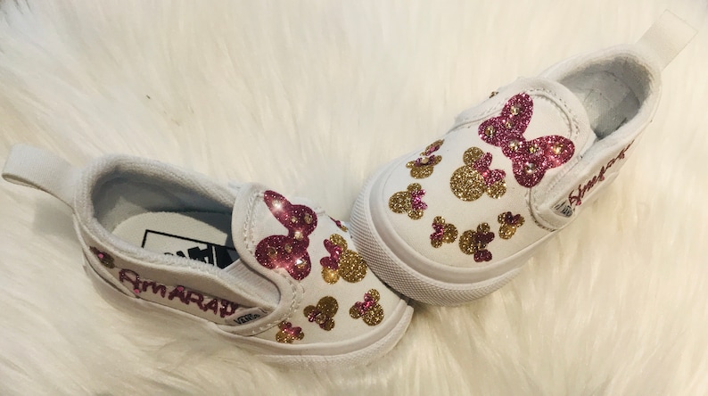 May include: A pair of white slip-on shoes decorated with pink and gold glitter. The shoes have a pink glitter bow and gold glitter Mickey Mouse heads. The left shoe has the name "Amaray" written on the side.