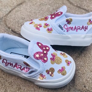 May include: A pair of light blue slip-on Vans shoes with gold and pink glitter Mickey Mouse designs. The shoes are personalized with the name "Amarah" in pink glitter.