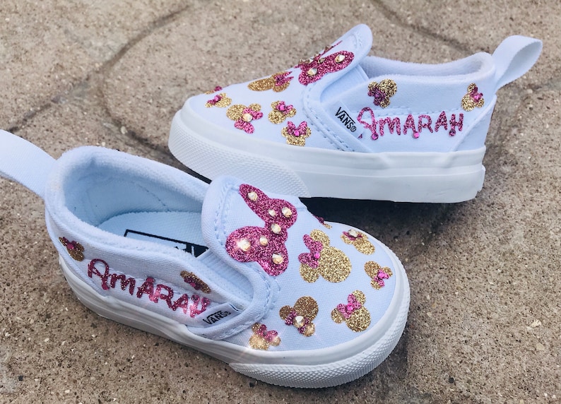 May include: A pair of light blue slip-on Vans shoes with gold and pink glitter Mickey Mouse designs. The shoes are personalized with the name "Amarah" in pink glitter.