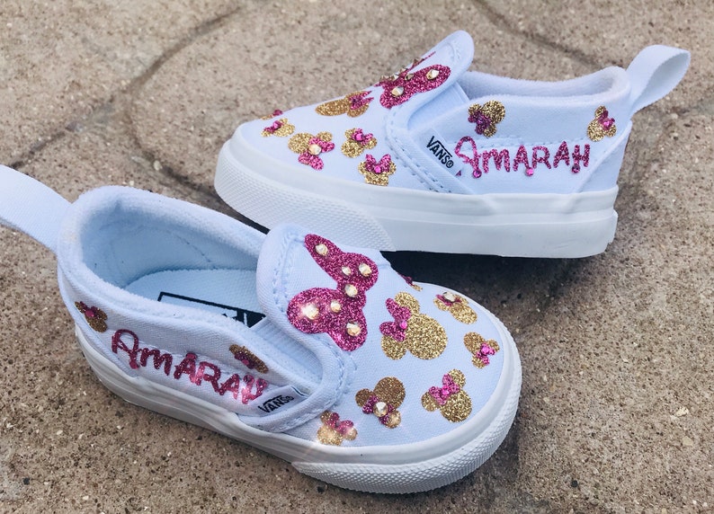 May include: A pair of light blue slip-on Vans shoes with gold and pink glitter Mickey Mouse designs. The shoes are personalized with the name "Amarah" in pink glitter.