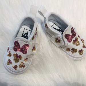 May include: A pair of white slip-on shoes with a pink bow and gold glitter Minnie Mouse designs.