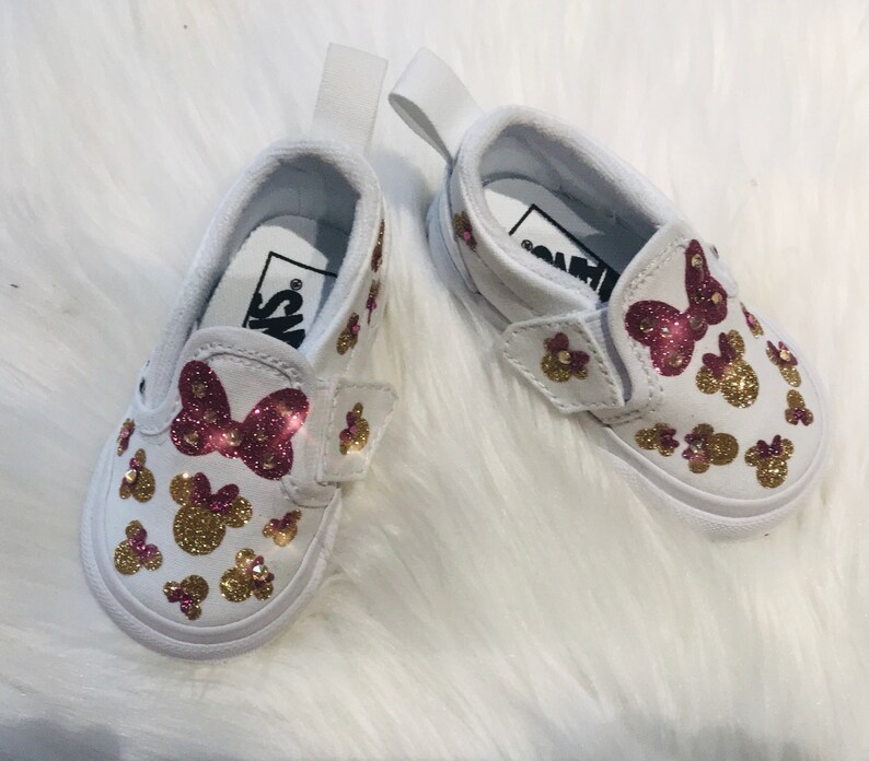 May include: A pair of white slip-on shoes with a pink bow and gold glitter Minnie Mouse designs.