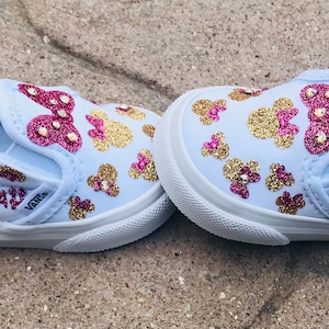 May include: A pair of white slip-on shoes with pink and gold glitter accents. The shoes have a Minnie Mouse design and the name "Amarah" is written on the side.