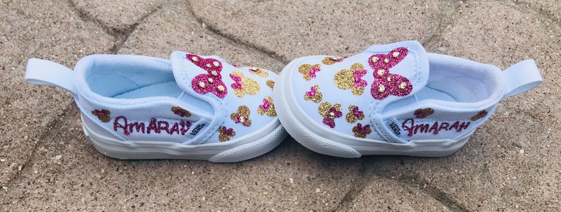 May include: A pair of white slip-on shoes with pink and gold glitter accents. The shoes have a Minnie Mouse design and the name "Amarah" is written on the side.