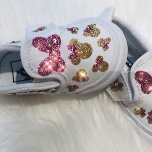 May include: White canvas slip-on shoes with pink and gold glitter Minnie Mouse designs. The shoes have a pink bow on the front and a velcro strap.