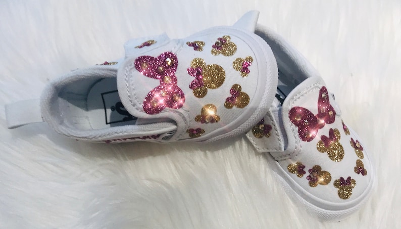 May include: White canvas slip-on shoes with pink and gold glitter Minnie Mouse designs. The shoes have a pink bow on the front and a velcro strap.