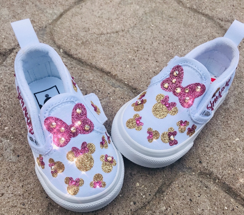 May include: White slip-on canvas shoes with pink and gold glitter Minnie Mouse designs. The shoes have the name "Shiloh" written on the side.