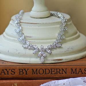 May include: A silver necklace with a teardrop-shaped crystal design. The necklace is on a white pedestal and a brown book with the text "Says By Modern Mast" on the cover.