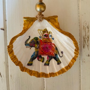 May include: A decorative seashell with a gold-painted edge and a painted elephant design. The elephant is adorned with colorful patterns and a red and pink blanket. A gold bead hangs from a jute string.