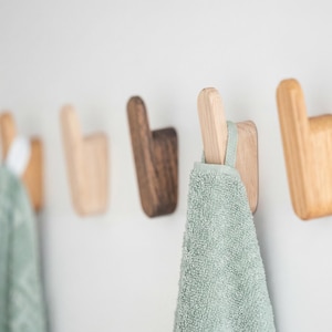May include: A series of wooden wall hooks in natural and dark wood tones. One hook holds a pale green towel. The hooks are mounted on a white wall, providing a minimalist and functional design for the home.