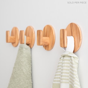 May include: Wooden wall hooks with a circular base and a rectangular hook. Two hooks hold light green towels, and one holds a white and green striped towel. The hooks are made of light-colored wood and are mounted on a white wall.