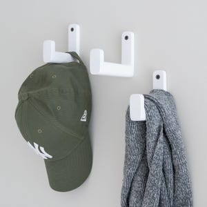 May include: Three white, L-shaped wall hooks. One hook holds a green baseball cap, and another holds a grey knit scarf. The hooks are mounted on a light grey wall, offering a minimalist storage solution.