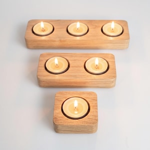 May include: Three light-coloured wooden candle holders with smooth, rounded edges. The top holder has three lit tea light candles, the middle holder has two lit tea light candles, and the bottom holder has one single lit tea light candle.