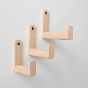 May include: Three wooden wall hooks in a light wood tone. The hooks are shaped like an L with a rounded end. They are mounted on a white wall.