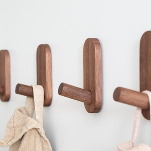 Könnte beinhalten: Ein Set aus vier Wandhaken aus dunkelbraunem Holz. Jeder Haken besteht aus einer vertikalen, abgerundeten rechteckigen Grundplatte mit einem hervorstehenden zylindrischen Stift. Zwei Haken halten helle Stoffgegenstände.