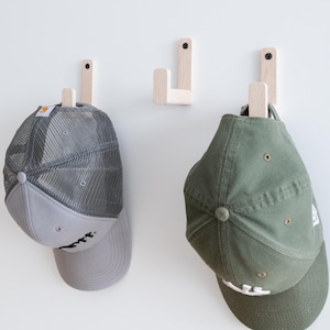 May include: Two wooden hooks mounted on a white wall with a gray and a green baseball cap hanging from them.