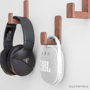 Puede incluir: Dos ganchos de pared de madera con unos auriculares inalámbricos negros colgados en uno y un altavoz portátil JBL blanco colgado en el otro. El texto "SE VENDE POR PIEZA" es visible en la parte inferior de la imagen.