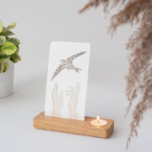 May include: A wooden candle holder with a small white candle in the center. A white card with a watercolor illustration of a bird flying above two hands is displayed in the holder.