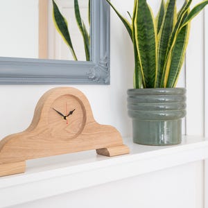 May include: A mantel clock crafted from light-toned wood with a smooth, arched silhouette. The clock face is integrated into the centre of the wooden frame and features black hour and minute hands, along with a thin red second hand.