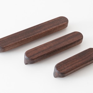 May include: Three dark brown wooden chopstick rests arranged diagonally. Each piece features a smooth, elongated rectangular shape with rounded ends and a visible wood grain pattern, showcasing a natural finish and a slightly curved underside.