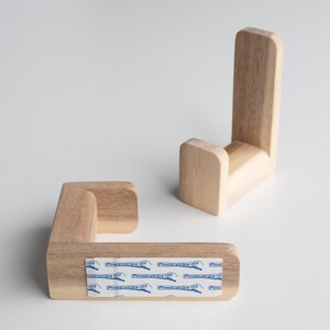 May include: Two wooden wall hooks with a J-shape design. The hooks are made of light brown wood and have a smooth finish. The hooks are attached to a white surface with adhesive strips labeled "Powersrips".