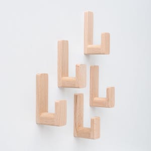 May include: Five light-colored wooden wall hooks arranged against a white background. Each hook has a rectangular base and a perpendicular, shorter arm, creating an L-shape. The hooks are made of natural wood.