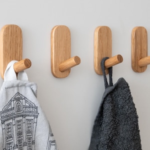 May include: Four light-colored wooden wall hooks with rounded rectangular bases and cylindrical pegs. A white fabric towel with a black architectural print hangs from the first hook, and a dark gray textured towel hangs from the third hook.