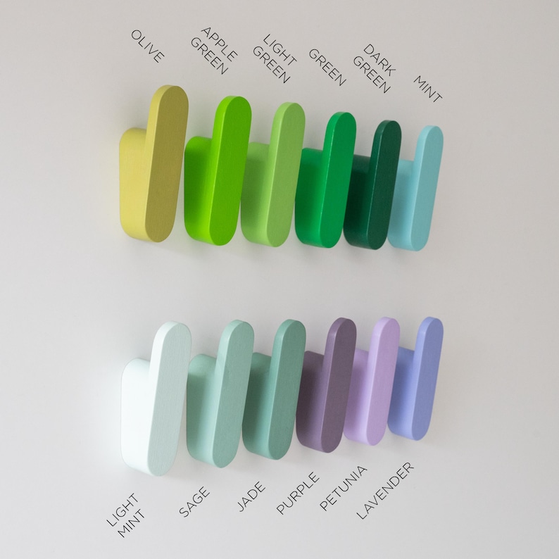 May include: A selection of wooden wall hooks in shades of green, purple, and blue. The hooks are labelled with colour names including "Olive," "Apple Green," and "Lavender." The hooks are mounted on a white wall.