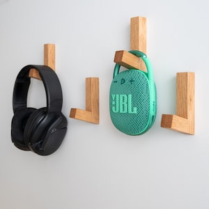 Op de afbeelding: Drie houten wandhaken met een zwarte over-ear koptelefoon en een groene JBL Clip 4 draagbare Bluetooth-speaker die eraan hangen.