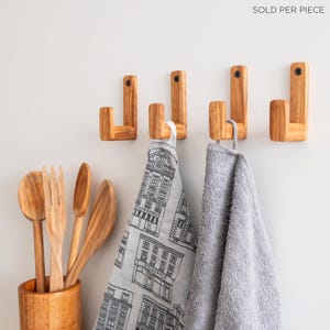 May include: Four wooden wall hooks with a rounded top and a square base. Two hooks have a gray towel hanging from them and one hook has a white tea towel with a black and white building pattern hanging from it.  The text "SOLD PER PIECE" is visible in the top right corner of the image.