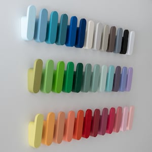 May include: Three rows of colourful wall hooks in various shades. The top row features blues, the middle row greens, and the bottom row oranges and reds. Each hook is a rounded rectangle shape, mounted on a white wall.