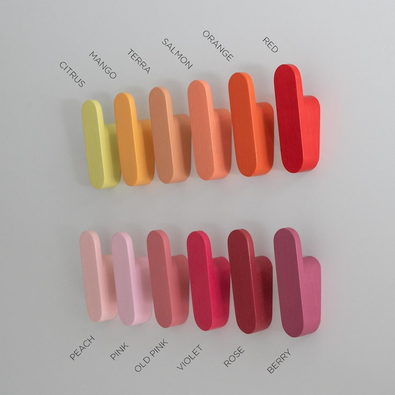 May include: A selection of wooden wall hooks in a range of colours, from citrus to berry. The hooks are labelled with their respective colours, including mango, terra, salmon, orange, and red. The hooks are mounted on a light grey wall.